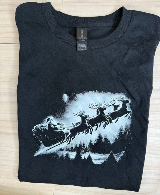 A black short-sleeve t-shirt with a white graphic print of Santa Claus in a sleigh pulled by reindeer over a snowy forest, laid flat on a light wood floor. The neck tag shows the brand as Gildan.