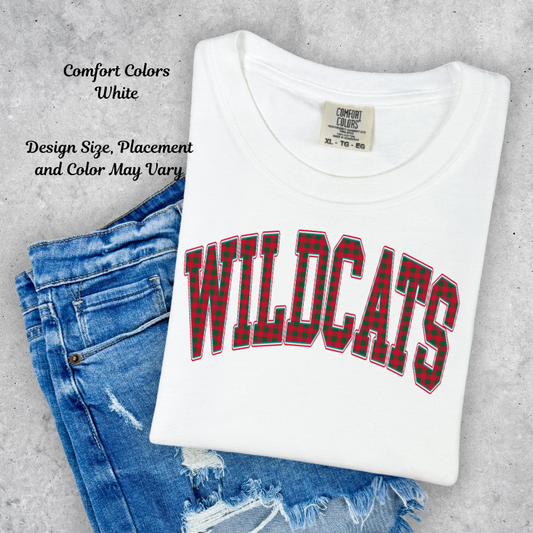 A folded white Comfort Colors t-shirt with 'WILDCATS' printed in red and black plaid block letters, styled on top of blue jeans against a gray concrete background. Text overlays on the left describe the shirt's color and sizing.