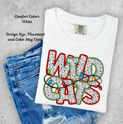 A folded white Comfort Colors t-shirt with a colorful graphic reading 'WILD CATS' in dotted letters, overlaid with Christmas string lights, styled on top of blue jeans against a gray concrete background. Text on the left reads 'Comfort Colors White' and 'Design Size, Placement and Color May Vary'.